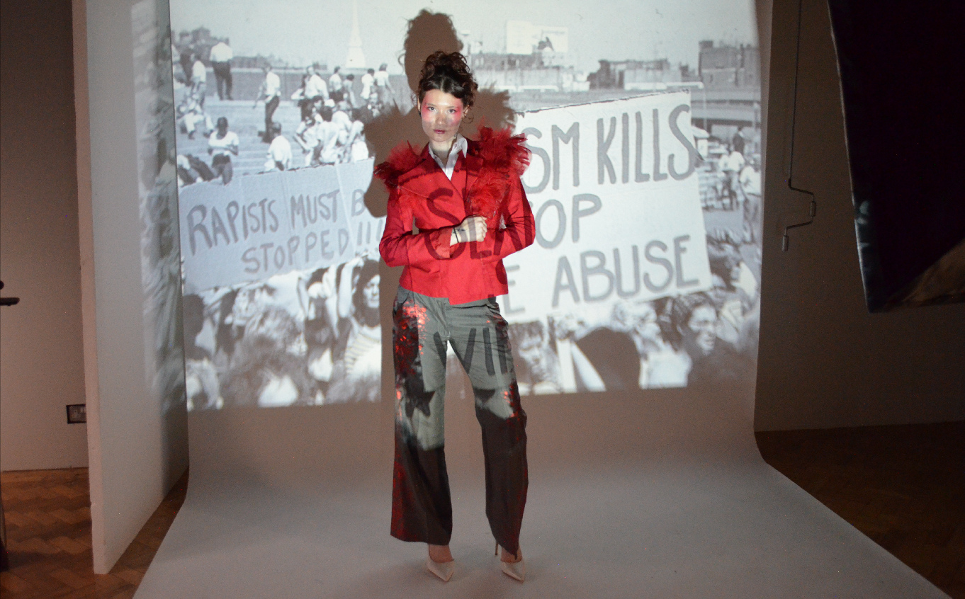 Female model wearing red top and grey trousers in front of anti-rape projection