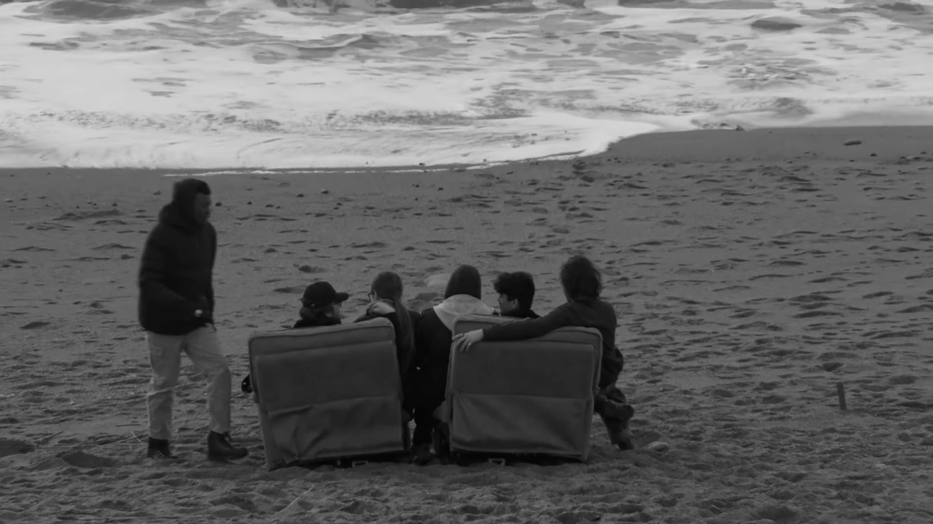 Black and white film still of group of people sitting on two lounge chairs on beach