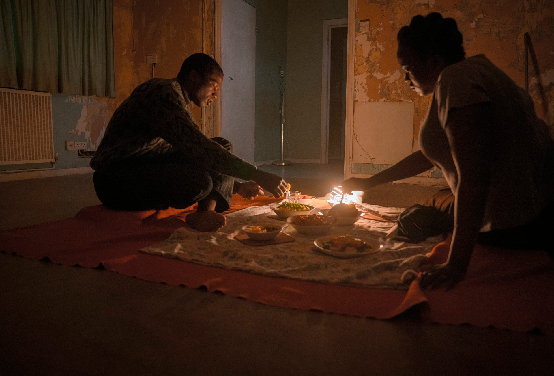 Still from 'His House' showing man and woman eating meal on blanket in undecorated room